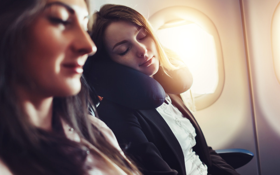 Eine Frau schl&auml;ft entspannt mit einem Nackenkissen im Flugzeug, w&auml;hrend die Sonne durch das Fenster scheint &ndash; ein Tipp zur Vermeidung von Jetlag.