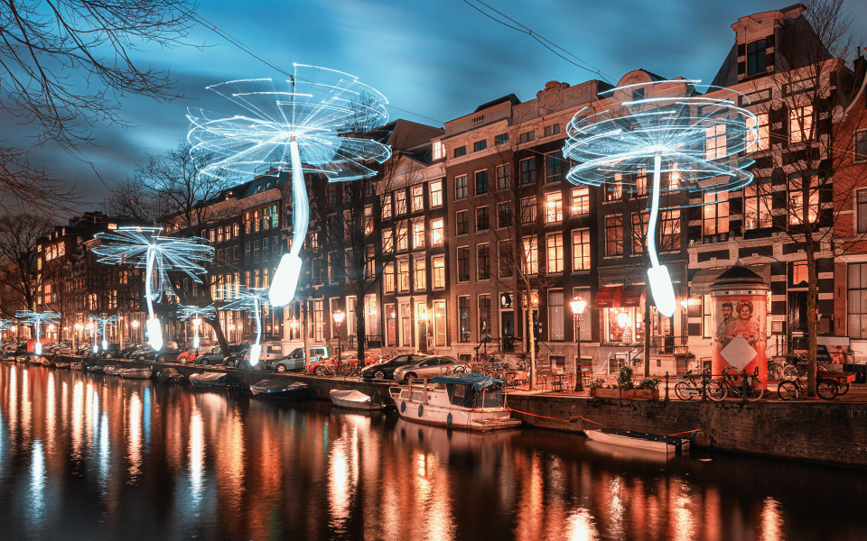 Leuchtende Kunstinstallationen in Form von Pusteblumen &uuml;ber einem Kanal in Amsterdam bei Nacht, mit reflektierenden Lichtern im Wasser und historischen Geb&auml;uden im Hintergrund.